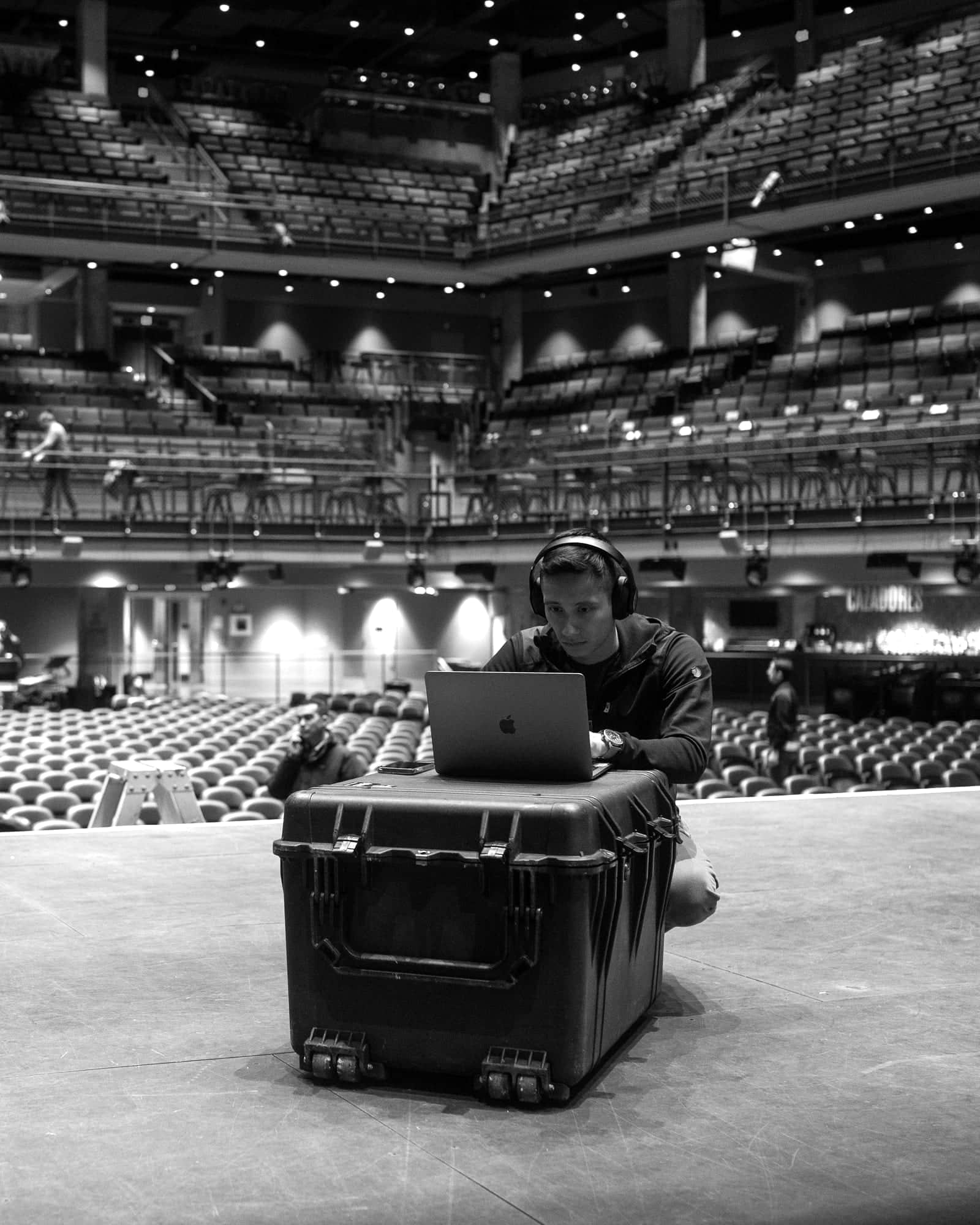 Ezra Zwaeli working on a laptop set on a road case at the edge of an empty theater stage, rows of seats rising behind him